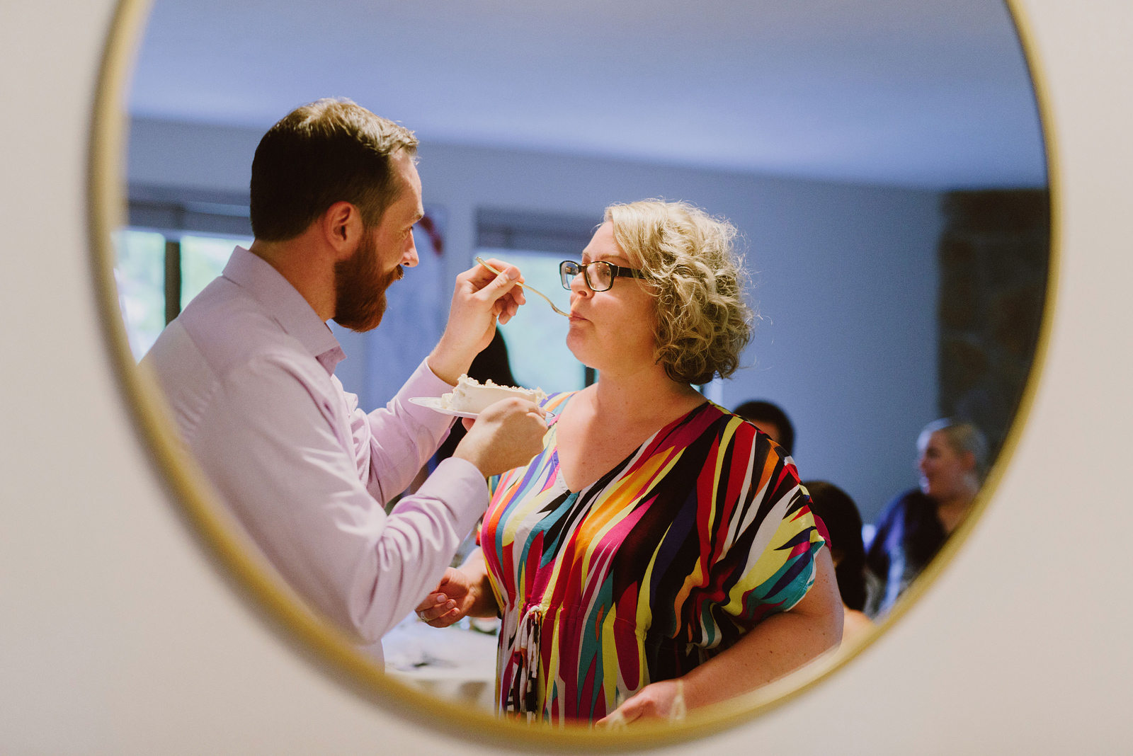 Candid photo shot through a round mirror of the groom feeding his bride a bite of cake - Oaks Pioneer Church Wedding