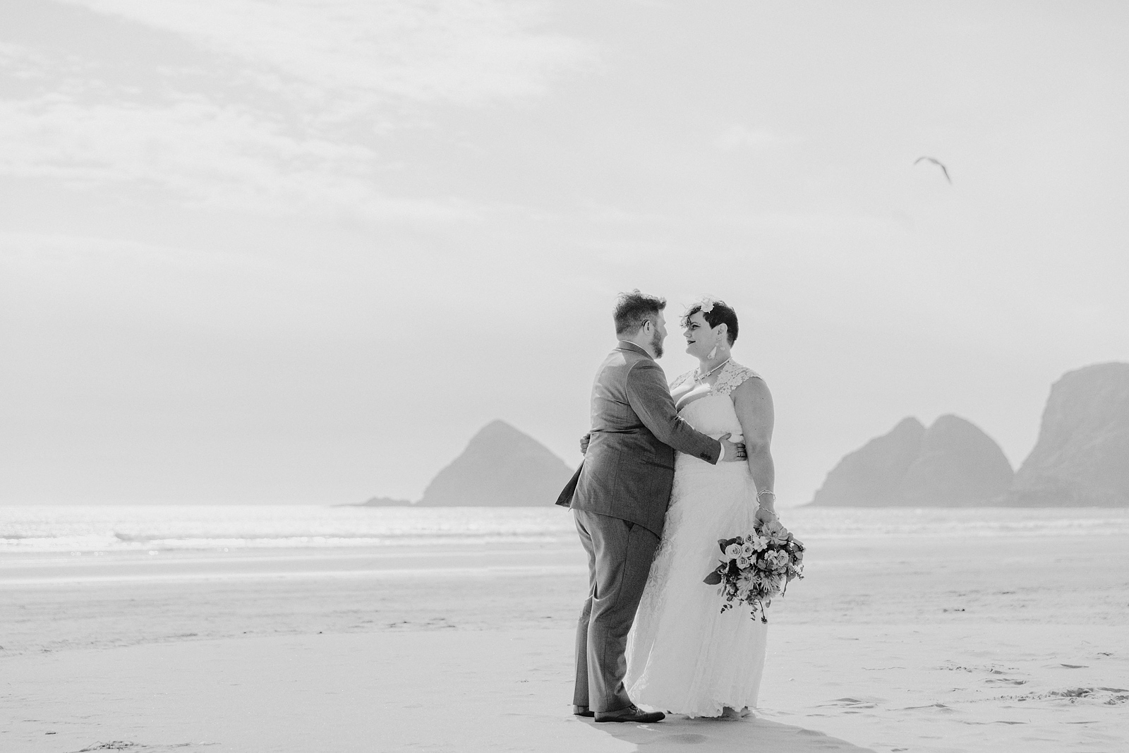 Black and white portrait of bride and groom embracing on the beach - Oceanside Community Club Wedding on the Oregon Coast” title=