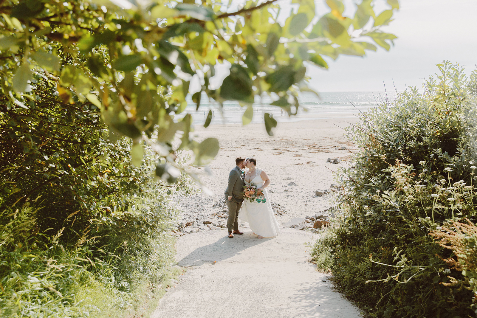 Bride and groom kissing on the beach under a branch of green leaves - Oceanside Community Club Wedding on the Oregon Coast” title=