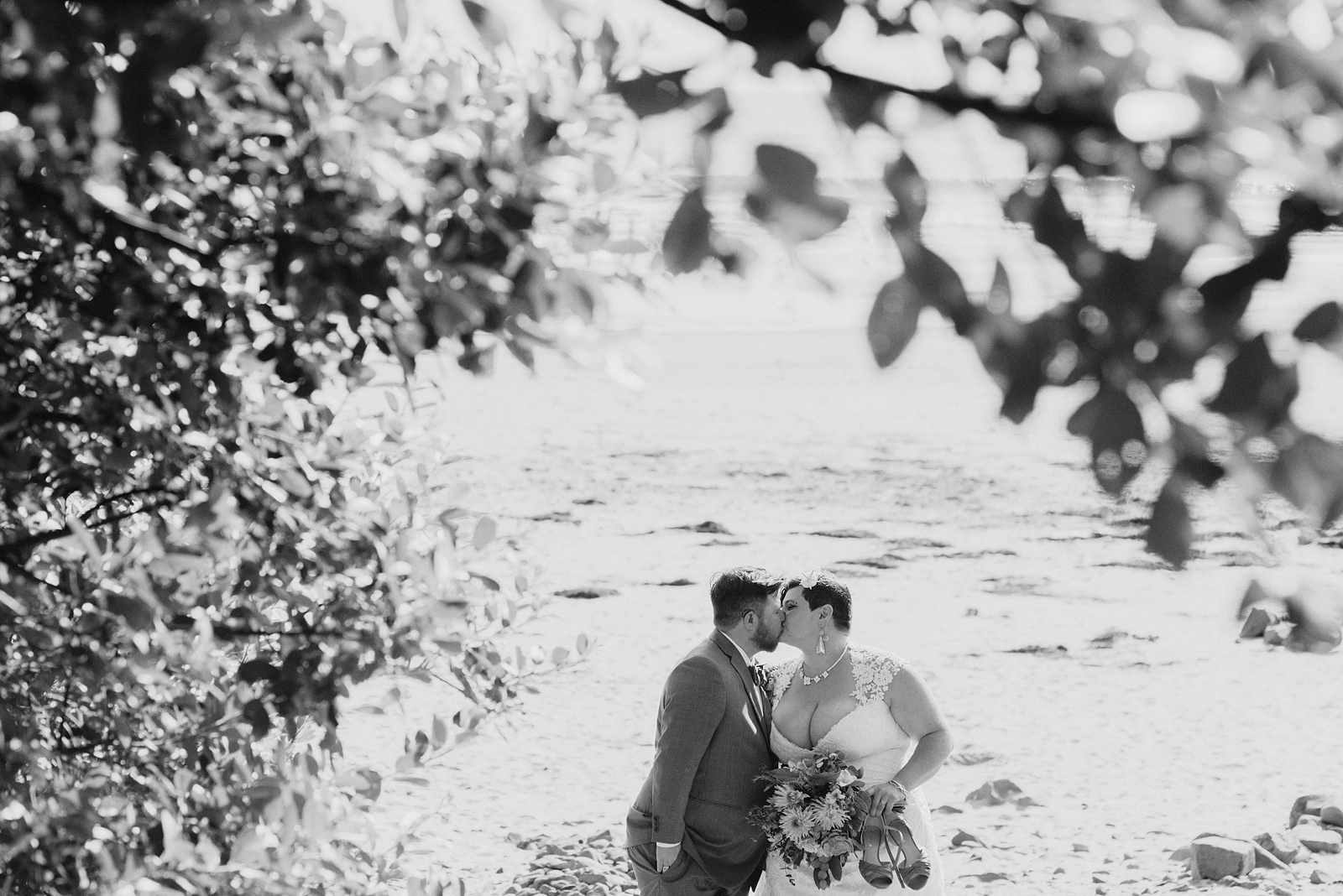 Black and white portrait of bride and groom kissing under a tree branch - Oceanside Community Club Wedding on the Oregon Coast” title=