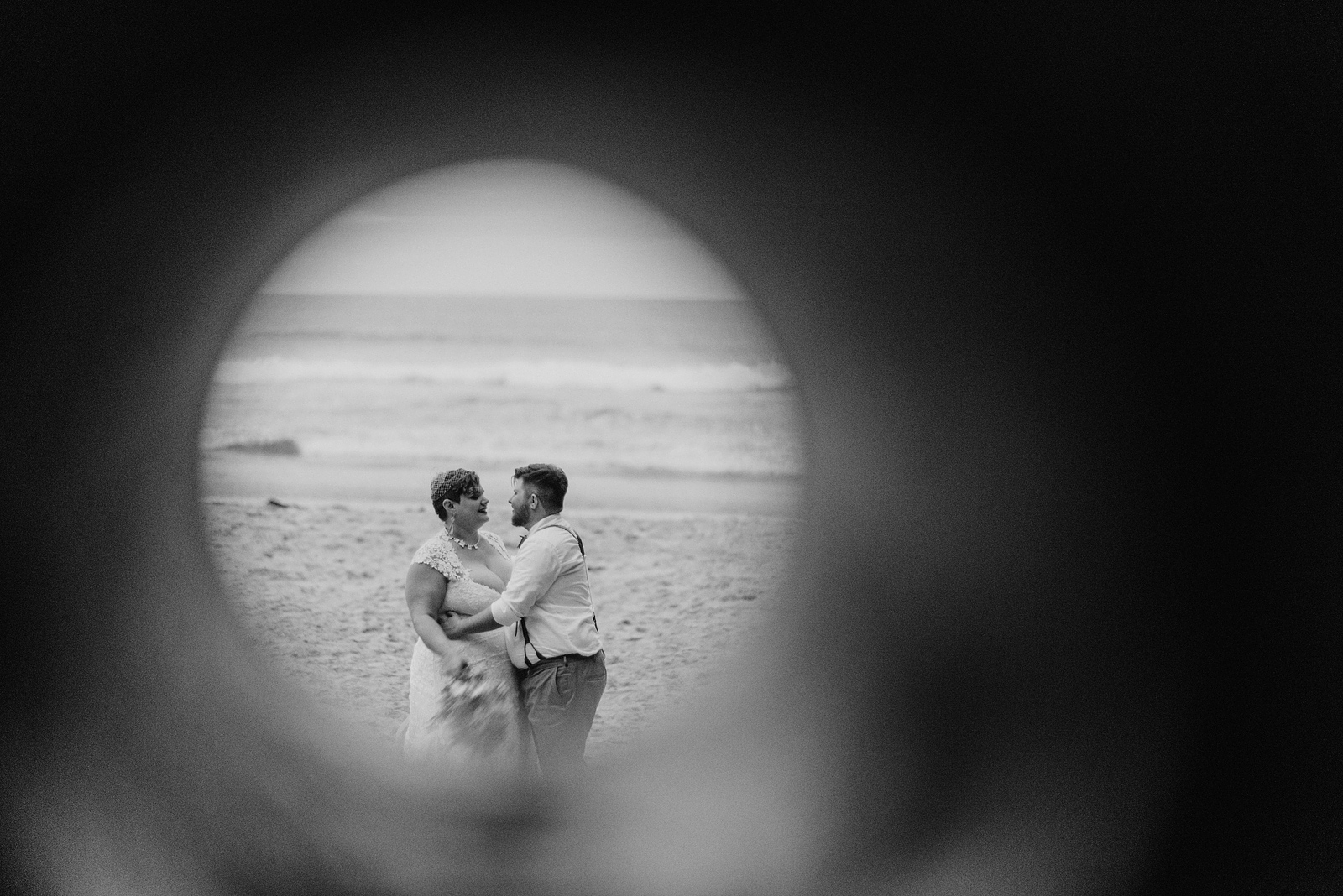 Creative black and white portrait of bride and groom taken through a hole in the wall - Oceanside Community Club Wedding” title=